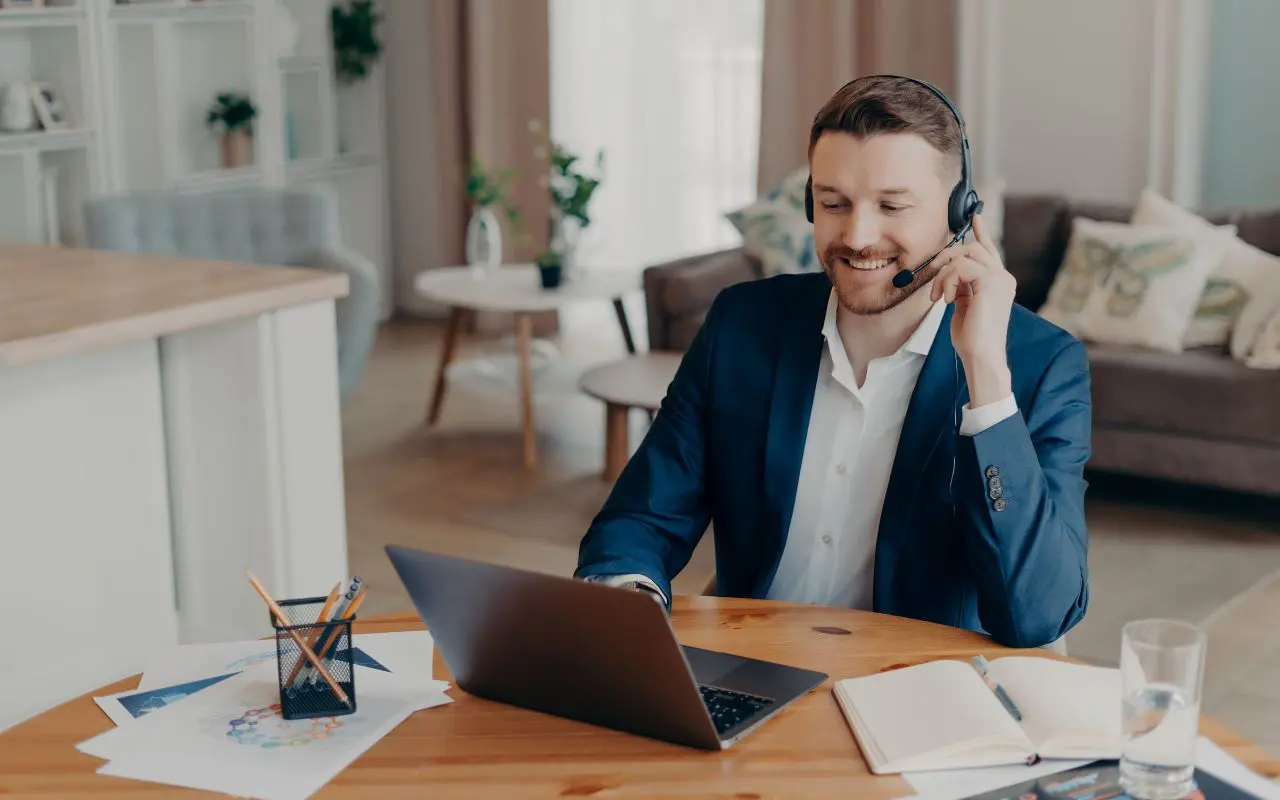 Photo d'un homme en blazer bleu avec un casque-micro souriant devant son ordinateur portable en télétravail