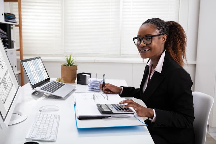 Photo d'une femme avec lunettes en tailleur noir utilisant une calculatrice à son bureau