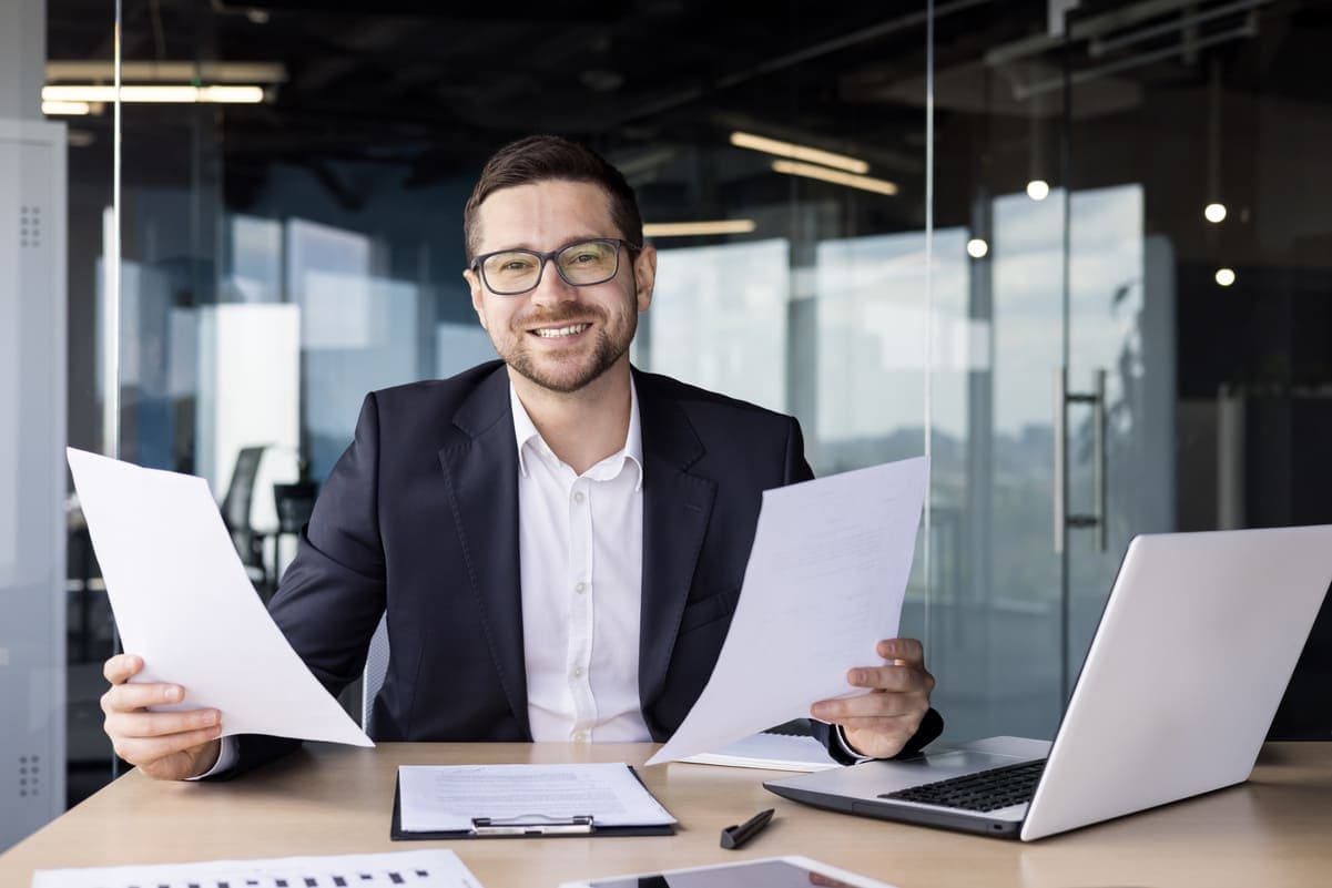 Photo d'un homme souriant en costume tenant des documents à son bureau avec ordinateur portable