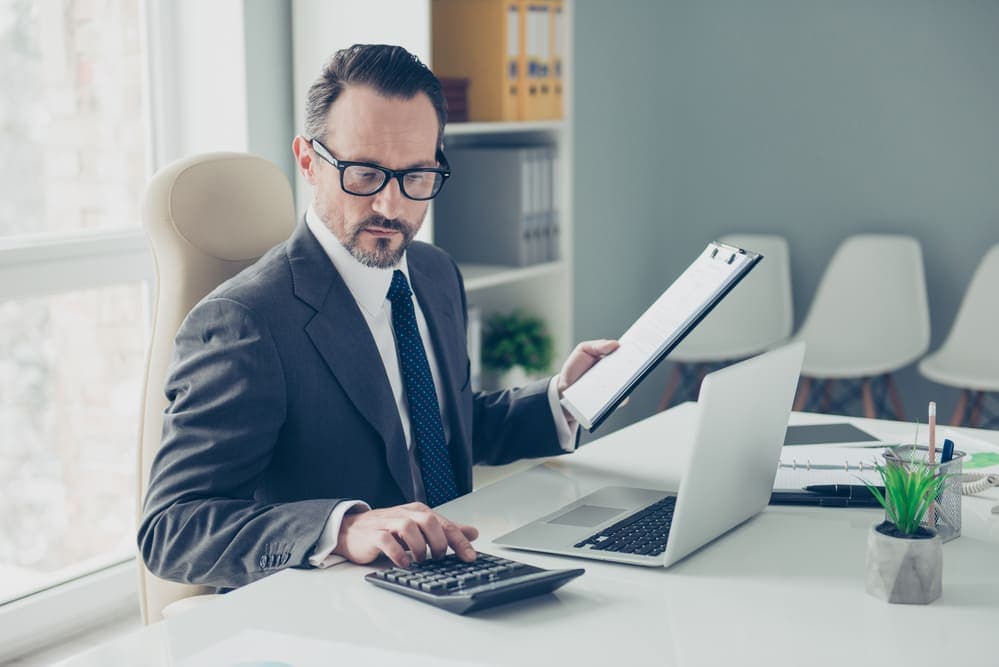 Directeur comptable vérifiant des documents financiers avec calculatrice et ordinateur portable à son bureau