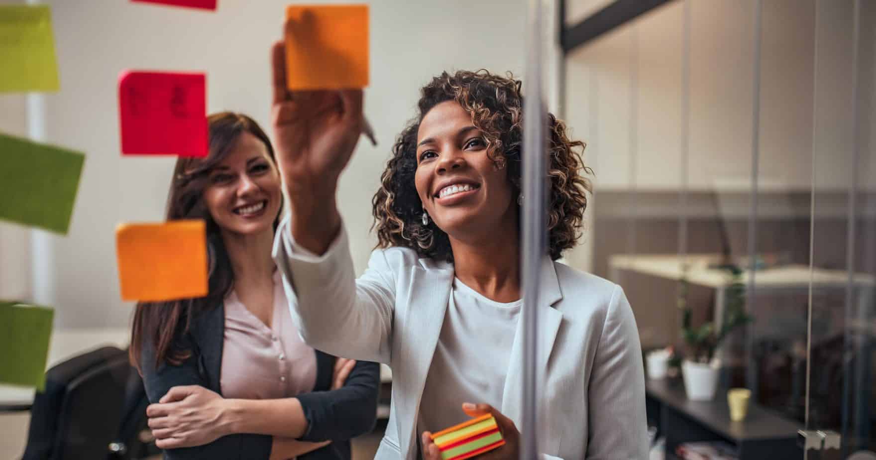 Photo de deux femmes collaborant avec des post-it colorés sur une paroi vitrée au bureau