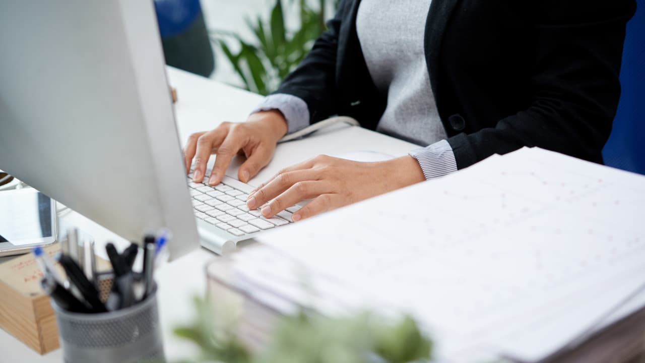 Photo de mains tapant sur un clavier devant un écran d'ordinateur avec agenda et stylos sur le bureau