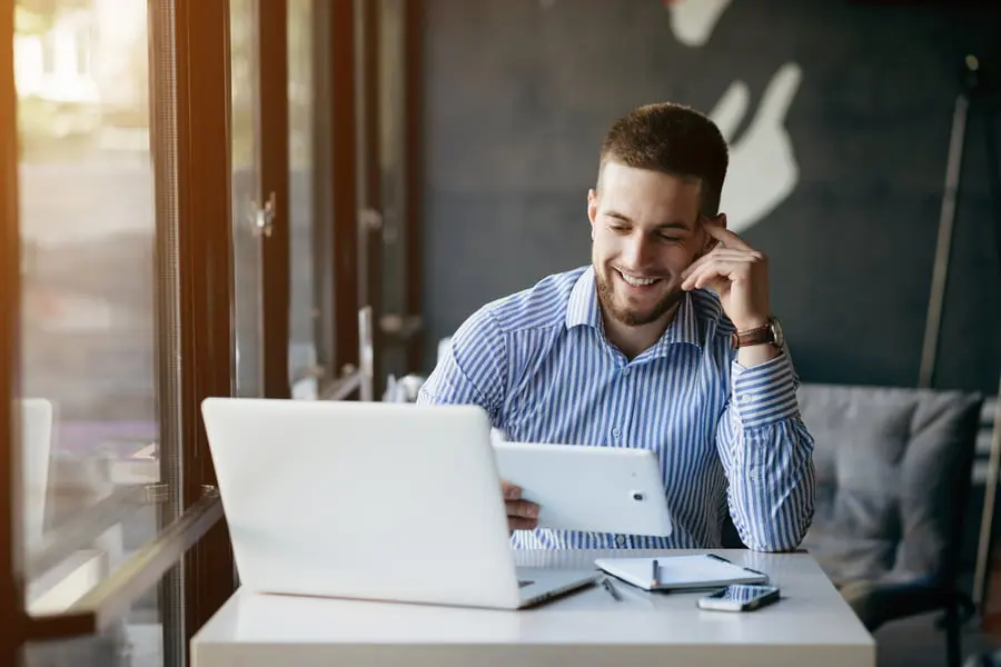 Photo d'un homme souriant en chemise rayée consultant une tablette devant son ordinateur portable