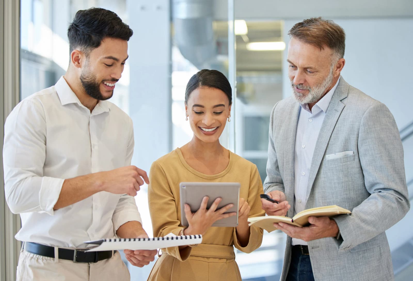 Photo de trois professionnels souriants consultant une tablette et des documents dans un couloir de bureau