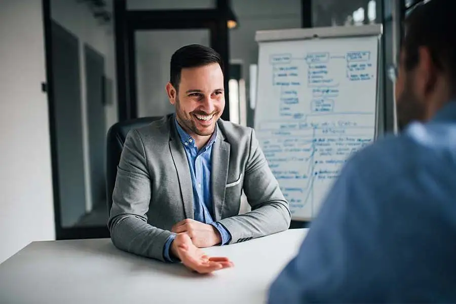Photo d'un homme souriant en blazer gris discutant avec un collègue devant un tableau blanc