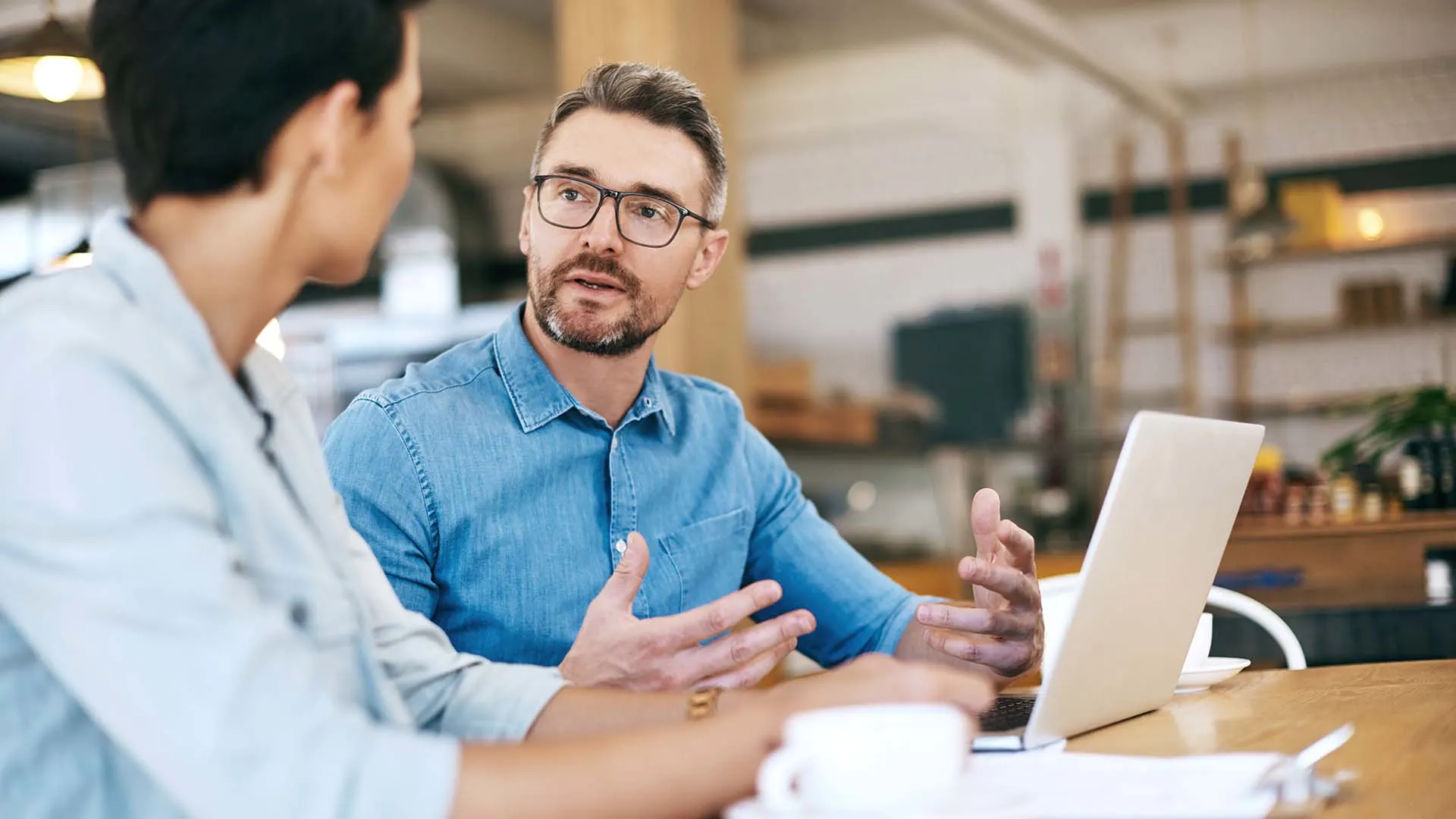 Photo d'un consultant en chemise bleue avec lunettes discutant avec un client devant un ordinateur portable