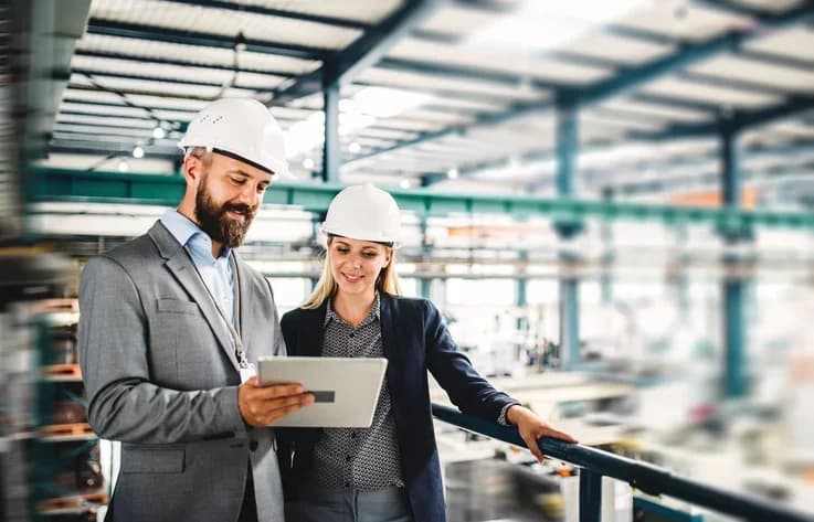 Photo de deux managers en casques consultant une tablette dans un hall industriel