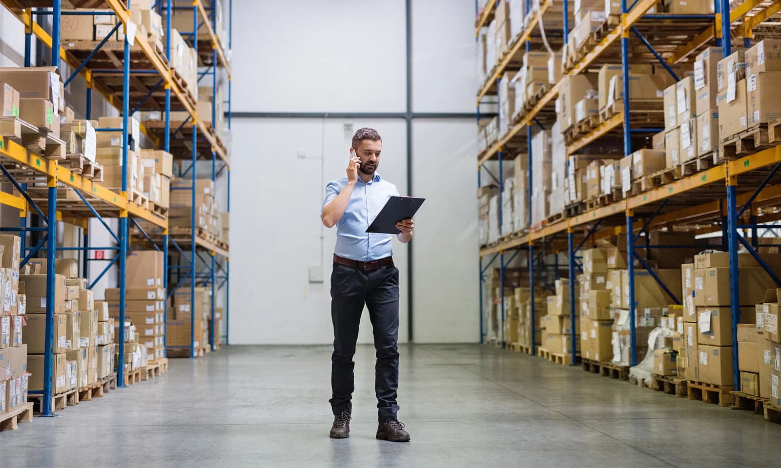 Photo d'un professionnel telephonant avec un dossier dans une allee d'entrepôt entre des rayonnages de cartons