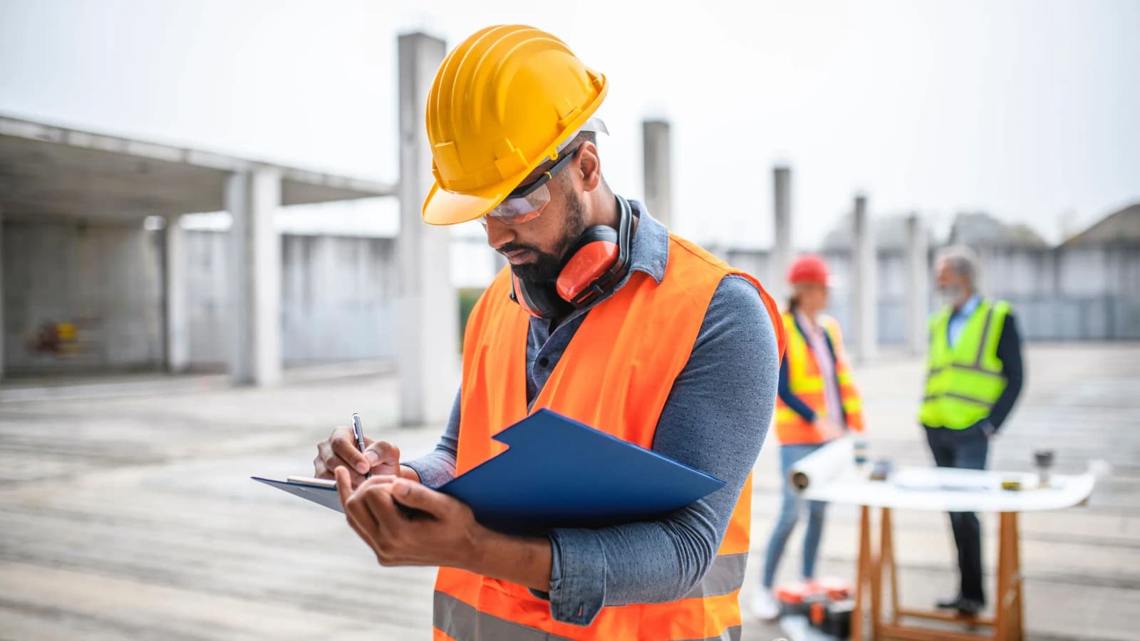 Photo d'un chef de chantier en casque jaune et gilet orange prenant des notes sur un porte-documents
