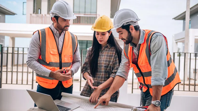 Photo de trois professionnels en casques et gilets orange etudiant des plans sur un chantier de construction
