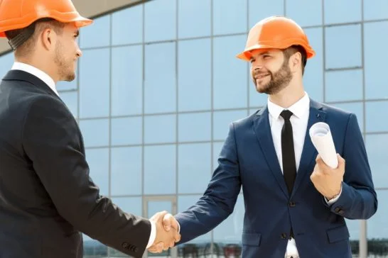 Photo de deux professionnels en costume et casque orange se serrant la main devant un bâtiment