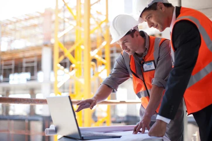 Photo de deux ingénieurs en gilets orange consultant un ordinateur portable et des plans sur un chantier
