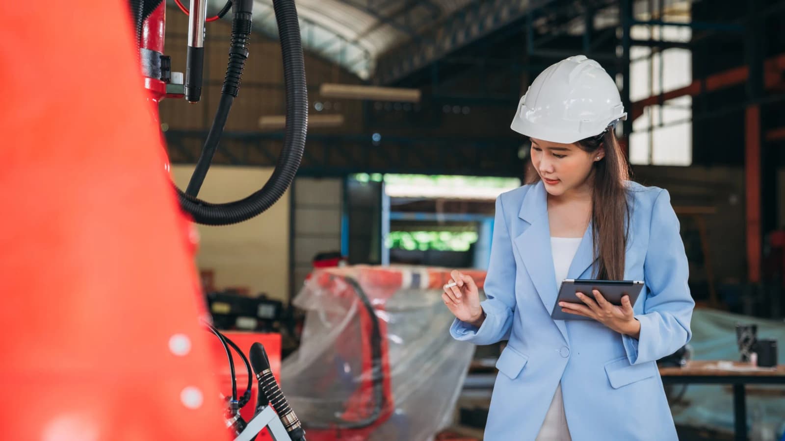 Photo d'une ingénieure en blazer bleu et casque blanc inspectant une machine industrielle rouge avec tablette