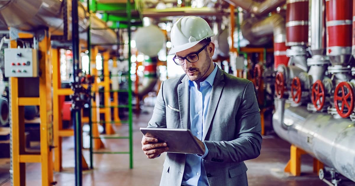 Photo d'un responsable en costume et casque blanc consultant une tablette dans une usine avec tuyauteries