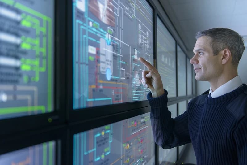 Photo d'un ingénieur pointant des schémas techniques sur un mur d'écrans de supervision