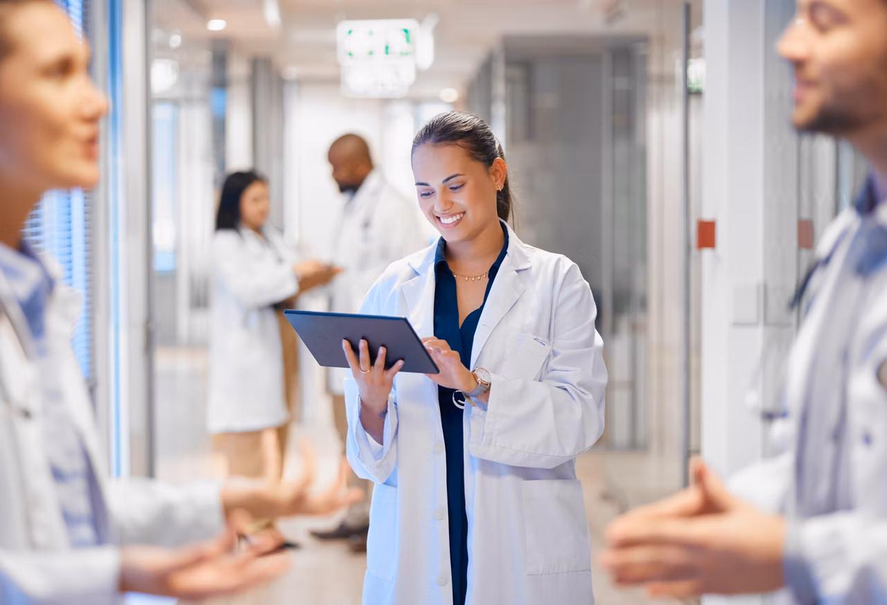 Cadre de santé en blouse blanche consultant une tablette numérique dans un couloir d'hôpital avec des collègues en arrière-plan