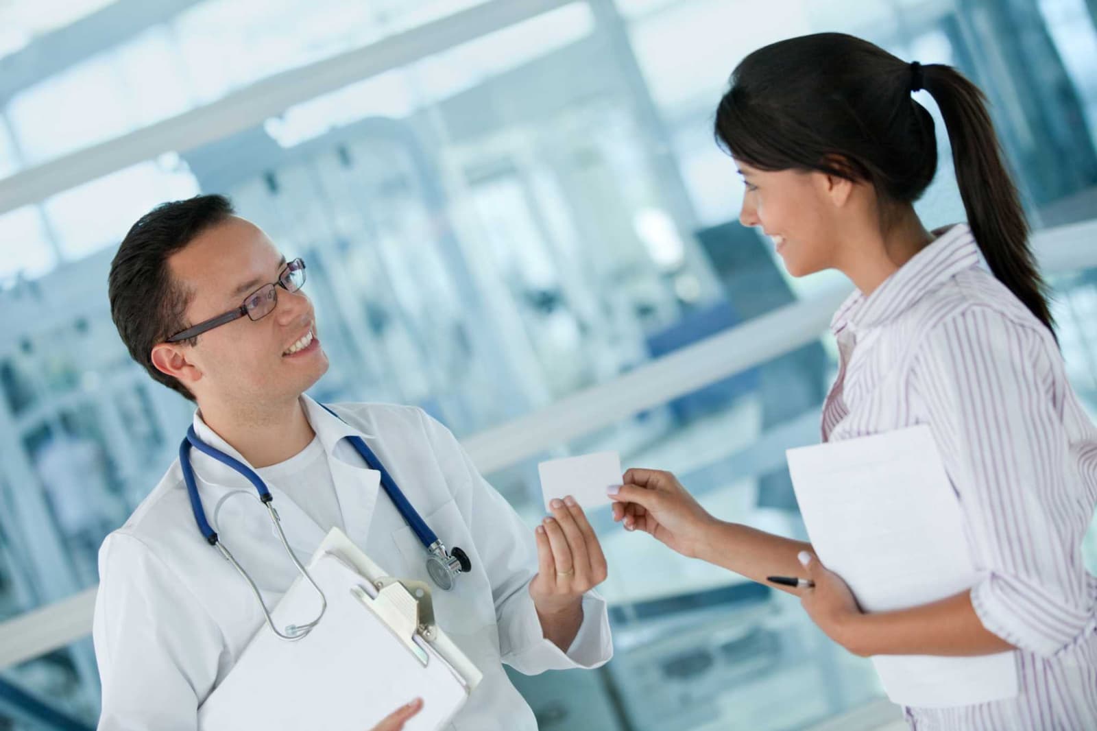 Photo d'un médecin en blouse blanche remettant une carte à une collaboratrice dans un hall vitré