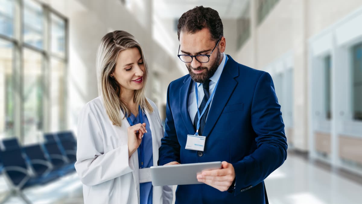 Photo d’un homme en costume et d’une femme en blouse blanche consultant une tablette dans un couloir